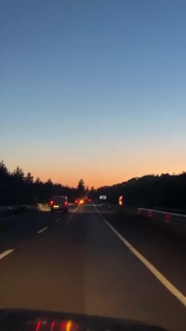 The video shows a car driving on a highway at dusk or dawn. The sky is a gradient of blue and orange. On either side of the highway are silhouetted trees. Several cars are ahead, their tail lights visible. The road markings are white and appear clear. The video is taken from the driver's perspective.