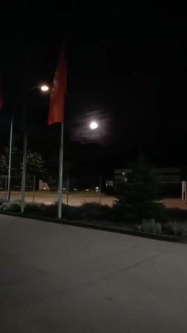 The video captures a serene night scene, likely in Lahr/Schwarzwald, Germany. The main subject is a bright, nearly full moon, prominently featured against a dark sky. The moon appears to be partially obscured by thin clouds, creating a luminous halo effect.

In the foreground, an orange flag hangs from a flagpole on the left side of the frame, illuminated by a nearby street light. The flag's design is not discernible. To the right, a dark evergreen tree stands out against the night. A paved road or pathway stretches across the bottom of the frame, with landscaped areas featuring shrubs and smaller trees separating it from a more distant background. In the distance, faint lights suggest buildings or structures.

The camera initially frames the scene with the flag and street light, then slowly pans upwards and slightly zooms in, bringing the moon into sharper focus as the central element. No people are visible, and there is no discernible activity other than the subtle camera movement. The time of day is clearly night. No text is visible in the video. The overall impression is one of quiet contemplation under a moonlit sky.