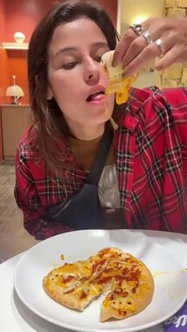 A woman wearing a red plaid shirt and multiple rings on her fingers is sitting at a white table with a white plate containing a piece of cheese pizza that has been partially eaten. She is using her right hand to lift a piece of the pizza towards her mouth. The pizza is melted with cheese and appears to have some red sauce and possibly bacon bits on top. The woman's mouth is open as she is about to take a bite, with a string of melted cheese stretching from the pizza to her mouth. She is wearing a dark bag across her chest. The background shows an indoor setting with orange and cream colored walls and decorative lighting fixtures.