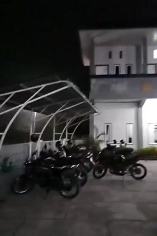 The media file shows a dimly lit outdoor scene at night, likely in Panteraya, Indonesia. The primary subject is a building with a modern, multi-story design, characterized by white walls, dark gray trim, and numerous windows and doorways. The building appears to be a lodging facility, possibly a hotel or guesthouse, indicated by a sign that reads "PENGINAPAN TUAH MANDIRI" (which translates to "Tuah Mandiri Lodging").

Several vehicles are visible in the foreground and on the property. These include motorcycles parked under a covered area, and various cars and a pickup truck parked in front of the building. Some of the vehicles are illuminated by the building's lights, while others are in shadow. Greenery, in the form of bushes or small trees, is present in planters around the building.

The time of day is clearly night, with the sky a deep black and artificial lighting from the building illuminating the scene. The weather appears calm, with no indications of rain or strong wind. The overall impression is one of a quiet, functional establishment operating into the evening. The lighting casts shadows and highlights the architectural features of the building, creating a somewhat stark but organized visual. There are no people visible in the media file. The emotions conveyed are neutral, focusing on the physical aspects of the location and its contents.