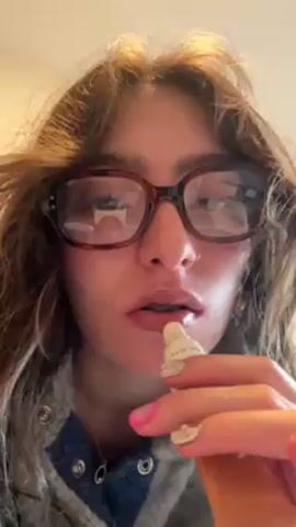 A young woman with medium-length brown hair and glasses is applying lip gloss. She wears a grey jacket with snaps and a silver necklace. She looks directly at the camera as she purses her lips. The background is blurry, showing a white ceiling.