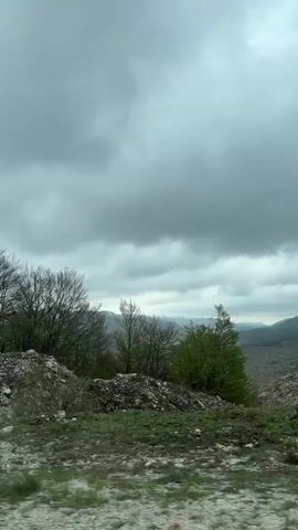 The video captures a panoramic view of a rugged, mountainous landscape in Provalija, Montenegro, under a heavily overcast, grey sky. The foreground features prominent, jagged rock formations and a ground covered in gravel and smaller rocks. Bare, deciduous trees, likely in late winter or early spring, are sparsely distributed across the rocky elevations. As the camera pans, it reveals distant hills and mountains, some with lighter patches suggesting remnants of snow or distinct geological features. A valley or lower-lying area with faint signs of greenery and scattered structures is visible between the foreground and the distant peaks. The overall scene depicts a cool, stark natural environment. No people or visible text are present.