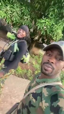 Two people are outside in a rural setting with trees. The person in the foreground is male, wearing a camouflage shirt and baseball cap. He is holding a phone and filming a selfie video. Behind him is another person, wearing a black shirt, a bandana, camouflage pants, and what looks like tactical gear. They appear to be smiling and walking together. The man in the foreground points upward at one point. In the background are rudimentary buildings constructed with sticks and grass.