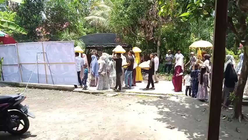 A group of people are gathered outdoors in a mostly unpaved area. They appear to be participating in some sort of procession or event. The majority of the people are dressed in traditional clothing, with many women wearing headscarves. Some individuals are carrying objects, including what appear to be large, yellow, umbrella-like structures. A white banner or screen is visible on the left side of the frame, supported by metal stands. In the foreground, a motorcycle is parked. The background consists of lush green trees and foliage, suggesting a tropical or subtropical environment. The overall atmosphere appears to be one of community gathering.