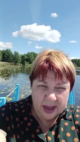 The video is a selfie-style recording of a woman with short, dyed blonde and red hair. She is wearing a dark green shirt with orange polka dots. She is outdoors with a body of water and trees in the background under a blue sky with clouds. She is looking at the camera and appears to be speaking, but there is no audio. The camera angle is from slightly below her face, looking up.