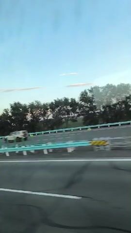 A view from a vehicle traveling on a highway. The sky is a clear blue with a few wispy clouds. Trees line the side of the road, separated from the highway by a green barrier. Cars and a truck are seen driving on the highway. The road is paved with asphalt and has white lane markings. The sun appears to be setting, casting a warm light on the scene.
