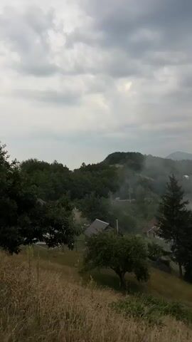 This media file depicts a serene, rural landscape under a cloudy sky. The scene is set in Mekića Kula, Montenegro, showcasing rolling green hills dotted with trees and small houses. The time of day appears to be late afternoon or early evening, as suggested by the muted light and overcast conditions.

The primary subjects are the natural elements of the landscape: the hills, the trees, and the sky. Several small houses with dark roofs are visible, blending into the natural environment. There are also some scattered objects that appear to be sheds or storage structures. Smoke or mist is rising from various points in the valley, adding a touch of mystery and tranquility to the scene.

The activity taking place is the peaceful existence of a rural community within its natural surroundings. The rising smoke could indicate local fires, possibly for cooking or heating, or it could be natural mist or fog rolling through the valleys.

Notable details include the dramatic, overcast sky with dark clouds that hint at impending rain or a storm. The lush green vegetation and the dry, golden grass in the foreground suggest a transitional season, possibly late summer or early autumn. The presence of the houses and smoke suggests human habitation and activity, but the overall feeling is one of quiet solitude and natural beauty. There is no visible text in the media file.