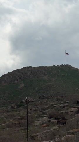 The video pans up from a ground-level view of a stony hillside with a simple wooden cross at the top, revealing a fortified structure atop a hill. The structure includes walls, a tower with a conical roof, and what appears to be an ancient fortress. People are visible on the hill, exploring the site. The surrounding environment includes a parking lot with numerous vehicles, a metal fence, utility poles with wires, and scattered vegetation. The sky is overcast, suggesting a cloudy day.