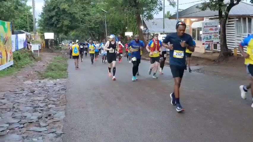 The video shows a marathon taking place on a paved road lined with trees and buildings. A group of runners, primarily men, are running towards the camera. They are wearing various athletic outfits, including shorts, tank tops, and t-shirts, with race numbers displayed on their chests. Many are wearing matching yellow and blue shirts. The road is damp, suggesting recent rain. On the left side of the road is a stone-covered area and a large billboard. On the right, there are low buildings with signs. The sky is overcast, indicating either early morning or late afternoon. The runners are spread across the road, with some individuals ahead of the main pack. The overall atmosphere is one of exertion and determination.
