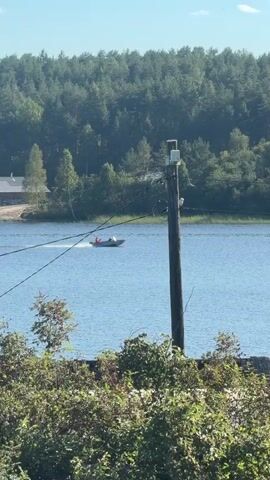 A boat with two occupants wearing orange life vests travels across a body of water. The boat is moving from left to right across the frame, creating a wake. In the background, a dense forest covers rolling hills, and a wooden utility pole stands near the water's edge, topped with a white rectangular apparatus. The foreground shows green foliage and a grey textured surface. The water is calm and reflects the sunlight.