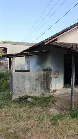 The video opens on a small, weathered house with a tiled roof and cracks in the stucco exterior. A green and white door is visible on the right side of the house, with a small covered porch. To the left of the house is a concrete structure, possibly a low wall. The ground in front of the house is covered with dry grass and some scattered rocks. Overhead, power lines crisscross the sky, which is a clear blue.

The camera then pans to the right, revealing a decorative gateway with blue and white pillars. Beyond the gateway, a paved path leads into what appears to be a village or residential area. There are trees and more houses in the background. A banner is hanging from a utility pole, with colorful graphics and text in Indonesian, advertising something related to children and education.

Further to the right, a cluster of lush green banana trees is prominent. The camera then shifts again to show a small building with a dark roof and blue double doors, possibly a shop or storage unit. A motorcycle with a black helmet resting on it is parked in front of this building. The surrounding area continues to be overgrown with grass. The lighting suggests it is either early morning or late afternoon, with the sun casting long shadows.