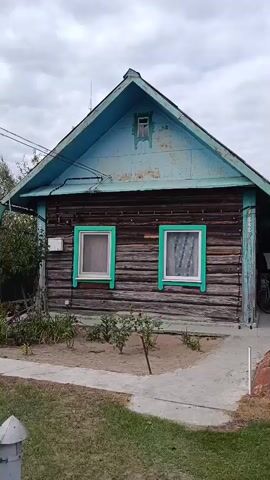 A static, medium shot video shows a weathered, log cabin with a blue, gabled roof. The cabin has two windows with bright green trim on the front facade, and a small, decorative window high up on the gable. The logs appear dark and aged, with some visible wear and tear. The roof is also painted blue, but the paint is peeling and faded. There is a small patch of garden in front of the cabin with some sparse greenery and a winding path leading up to the entrance. The sky is overcast and appears cloudy. To the right of the cabin, a bicycle is partially visible. The overall scene suggests a rural or rustic setting.