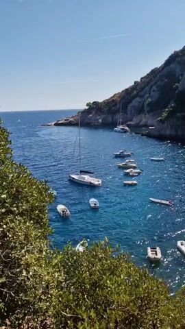 The video shows a serene bay with clear blue water. Several boats are anchored in the bay, including a large sailboat, a catamaran, and several smaller motorboats and dinghies. The bay is surrounded by a rocky, green hillside. The sun is shining, and the water is sparkling. The scene appears peaceful and idyllic.