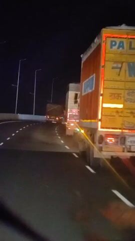 The video shows a highway at night with multiple trucks on the road. The perspective appears to be from inside a vehicle, likely another truck, as the camera is positioned low and close to the road. Streetlights illuminate the dark sky and the highway ahead. The trucks are large cargo vehicles, with their taillights and reflective markings visible. One truck in the foreground is particularly prominent, showing its rear and side. The road has white lane markings, indicating a curved path. The overall scene depicts a nighttime journey on a highway, with the heavy traffic of trucks suggesting commercial transport.