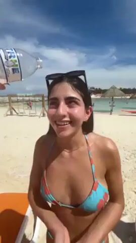 A young woman in a floral bikini stands on a sandy beach. Someone off-camera pours water from a plastic bottle over her head. She closes her eyes, smiles, and laughs as the water runs down her face. Sunglasses are perched on top of her head. In the background are other people, various beach structures, and the ocean under a partly cloudy sky.