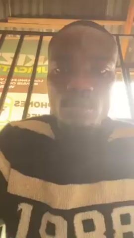 A close-up shot shows a man with dark skin wearing a black and beige striped sweatshirt with "1986" printed on it. He is indoors, standing behind a metal grill. His head is shaved, and he has a slight mustache. The background shows a sign with the word "KITCHEN" visible and assorted items on shelves, suggesting a shop or stall. The camera angle is slightly upward, giving the viewer the impression of looking up at the man. He glances to the side with a slightly intense expression.