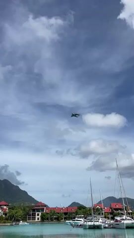 A military jet flies across a cloudy blue sky above a harbor with sailboats docked. In the background, there are buildings and lush green mountains.