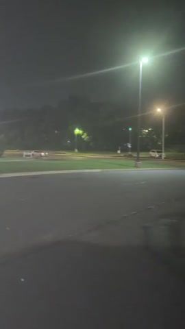The video shows an outdoor scene at night. Several streetlights illuminate a paved area, possibly a parking lot or road, and a grassy hill. A few cars are visible, one white car in the left foreground appears to be turning. Another white car and a darker vehicle are further in the background. Several green-capped lights are spaced along the edge of the grassy area. A red stop sign is also visible on the grass. Trees form a dark backdrop against the night sky. The lighting from the streetlights creates strong glare and shadows.