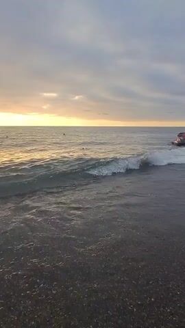 The video shows a beach with waves crashing on the shore. The sky is cloudy with the sun setting in the distance, casting a golden glow on the water. In the distance, a jet ski is visible on the right side of the frame. The foreground of the video shows the beach with small pebbles.