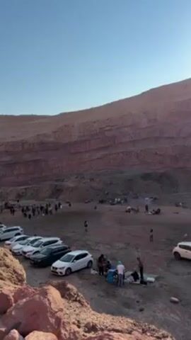 The video shows a large group of people gathered in a quarry, seemingly for an outdoor event or gathering. The quarry has layered, reddish-brown walls and a flat, dirt-covered floor where the people are congregated. Several cars are parked along one side, near some large rocks. Groups of people are scattered across the quarry floor; some appear to be setting up food or sitting around, while others are walking around. The sky is clear and blue. Upbeat music is playing.