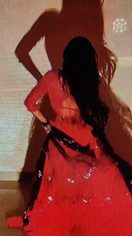 A person dances in a red outfit. The person has long black hair and is wearing a flowing red dress with a black accent. The background is a textured wall with a shadow of the person projected on it. The lighting is dramatic, casting shadows and highlighting the person's movements.