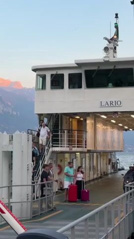The video shows a ferry docked, with passengers disembarking. A barrier arm with red and white stripes and illuminated red lights is visible on the left. People are walking down stairs from the upper deck and on the main deck of the ferry, some are carrying luggage. A man in a suit is walking with two people carrying red and pink suitcases. A person on a scooter is riding past them. The ferry is named "LARIO". In the background, there is a body of water and mountains with a sunset glow.