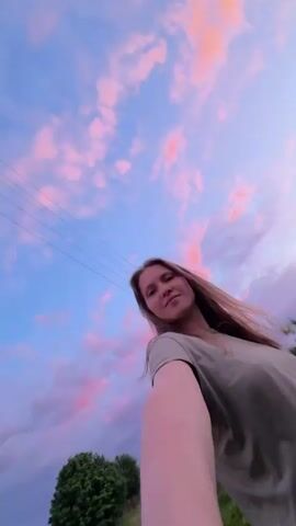 A young woman is taking a selfie outdoors at sunset. She is wearing a green t-shirt and has long brown hair. The sky is filled with colorful clouds, with shades of pink, orange, and blue. In the background, there are trees, a fence, and some greenery. Power lines are visible in the sky. The camera angle is low and tilted upwards, capturing the woman and the sky. The woman smiles at the camera.