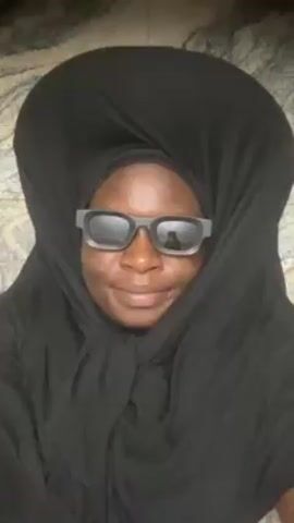 A person wearing a black headscarf and sunglasses puckers their lips towards the camera. The sunglasses have thick, gray frames. The person has dark skin. The background is a blurry, light gray surface, possibly granite. The person appears to be indoors. There's a reflection of a person holding a phone in the lens of the sunglasses.