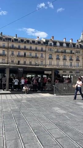 The video shows a wide-angle view of a busy city square on a sunny day with scattered clouds. In the foreground, a patterned gray stone plaza is traversed by pedestrians, including a woman walking diagonally across the frame. A black tram stands at a station, with people waiting to board. The background features ornate, multi-story buildings in a classical architectural style, some with signage. A tall, modern lighting structure stands adjacent to the tram line. The overall impression is a bustling urban scene in a European city.