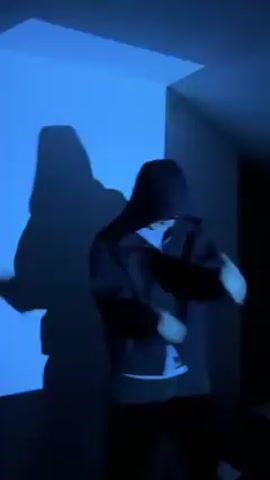 The video shows a person dancing in a room lit with red light. The person is wearing a dark jacket, sunglasses, and has the hood up. They are dancing and making hand gestures. The red lighting creates a stark contrast between light and shadow, emphasizing the silhouette of the figure and casting a large shadow on the wall behind them. The walls of the room are visible, creating a sense of enclosure. The music is audible, suggesting a performance or music video setting.