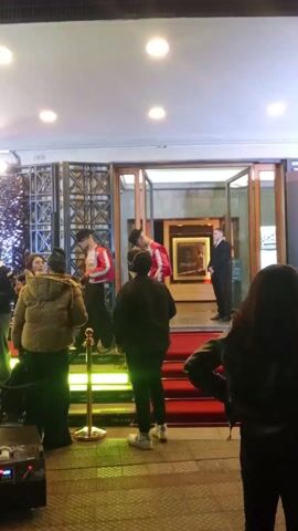 At an outdoor evening event, two individuals dressed as Michael Jackson walk a red carpet. They wear red jackets, black pants, and hats. They are surrounded by people, some of whom are taking pictures. A banner reads “Michael” and “23 de Abril SOLO EN CINES”. A black sequined wall is in the background. The entrance to a building is visible behind the red carpet, with a security guard standing inside. There are yellow rope barriers separating the people from the red carpet.