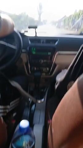 The video shows the interior of a car driving on a paved road. The camera is positioned behind the front passenger seat, looking forward. In the driver's seat, a person's arm is visible on the steering wheel. The dashboard displays a radio and gear shifter. The rearview mirror reflects the road ahead and the driver's face. Outside the car, trees and buildings line the road, with other vehicles visible in the distance. The lighting suggests it is daytime.