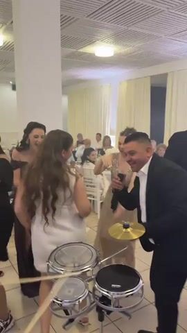 The video captures a lively wedding reception scene. A man in a suit with an open white shirt sings into a microphone while standing near a small drum set.  A woman in a white dress, holding drumsticks, dances near the drums with another woman in a glittery gold dress. Other people are dancing and clapping around them, including a man in a tuxedo who briefly holds a saxophone. In the background, guests are seated at tables with white linens. The room has white walls, white draperies, and a modern ceiling design with recessed lighting. The mood seems festive.