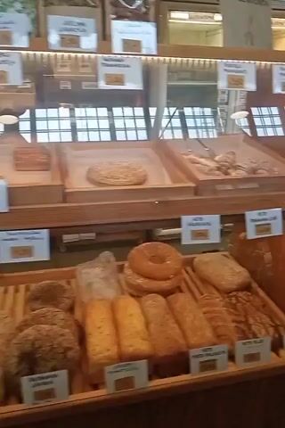 This is a short video of a bakery display case filled with various baked goods. The display case is made of wood and glass. On the top shelf, there are croissants and some other pastries. The middle shelf has a large ring-shaped bread and a loaf of bread with a dark crust. The bottom shelf features several loaves of bread, some of which are sliced. There are also small signs with prices and names of the products in Finnish. The lighting in the bakery appears warm and inviting. The video is shaky, suggesting it was filmed by someone holding a phone or camera. There are no people visible in the frame, only the baked goods. The overall impression is that of a well-stocked and appealing bakery.