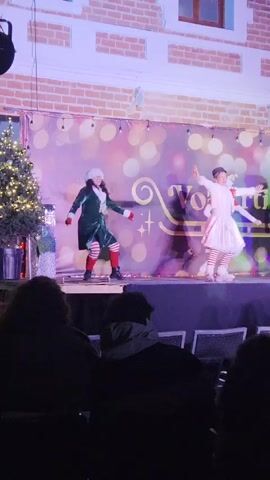 Here's a description of the video:

A performance featuring multiple dancers on a stage. They are dressed in Christmas-themed costumes: elf-like outfits in colors like red, green, white, and some with striped stockings. The stage backdrop is decorated with bokeh lights and text. The stage is lit with purple-toned lighting.

The dancers are actively moving, seemingly performing a choreographed routine. They wave their arms and step in sync.

The audience is visible in the foreground, sitting in chairs and watching the performance. Some members of the audience are holding up their phones, possibly recording the show.

The performance is taking place outdoors. A brick building is visible in the background behind the stage. A small Christmas tree is visible to the left side of the stage.