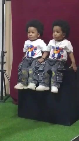 The video features two young children, likely twins, sitting side-by-side on a dark, cuboid prop. Both children have dark, curly hair and are dressed identically in white t-shirts adorned with a cartoon bear wearing sunglasses and a baseball cap. They are also wearing distressed denim-style cargo pants with gold-colored patterns and white sneakers.

The child on the left is visibly smiling and appears cheerful, occasionally kicking their legs. The child on the right maintains a more neutral or curious expression, looking slightly to their right.

The setting is an indoor space with a plain dark red or maroon backdrop and a green artificial grass surface on the floor. A black tripod stand is visible on the left side of the frame, suggesting a photo or video shoot setup. An adult's voice can be heard off-camera throughout the clip, seemingly interacting with the children, accompanied by background conversation and lighthearted sounds. The overall atmosphere appears to be a casual children's portrait session.