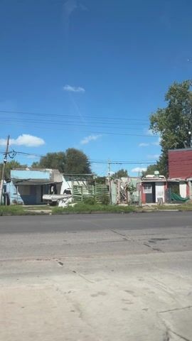 The video shows a view from a moving vehicle driving along a paved road on a sunny day. The sky is bright blue with scattered clouds. On the side of the road, there are several small, rundown buildings of various colors, including red, white, blue, and green. Some of the buildings appear to be residences, while others might be small shops or businesses. There are trees interspersed among the buildings, and power lines run above the street. As the vehicle moves, a cyclist and a motorcyclist pass by on the road. The general impression is a low-income urban or suburban area.