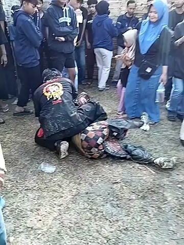 A medium shot depicts a group of people gathered outdoors. In the foreground, two figures, dressed in black clothing, are on the ground. One figure is kneeling over the other, who is lying face down. Both are splattered with a red substance resembling blood. A crowd of onlookers surrounds them, some with arms crossed. The ground is covered with sparse vegetation and bits of debris.
