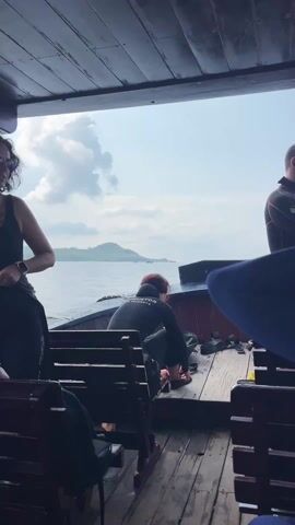 The video depicts several individuals on the wooden deck of a boat, seemingly preparing for or participating in a water activity in the Natural Heritage Area Trang, Vietnam. The scene is set at sea, with distant islands or hills visible under an overcast sky.

Multiple people are present on the boat. One woman, wearing a black tank top and sunglasses on her head, is seen adjusting her hair. Another woman, identified by the text "INSTRUCTOR Julia Vorobyeva" on her black wetsuit, is crouched on the boat deck, appearing to be arranging equipment. A man in a black wetsuit with red accents, marked "CRESSI," is also seen on deck, looking down at something in his hands. Another man in a black jacket stands beside him, looking out towards the sea. In the final moments of the video, a woman in a blue wetsuit is shown from a closer perspective, adjusting her hair at the back of her neck.

The boat's interior features wooden benches and a wooden deck. The general atmosphere suggests preparation for a dive or snorkel excursion, given the wetsuits and the instructor's presence. The weather is bright but cloudy, and the sea appears calm.