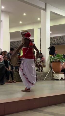 A man is dancing and playing a small drum on an indoor stage in front of a seated audience. The man is wearing a white turban, a red and white outfit, and is barefoot. He is facing away from the camera. He is hitting the drum with both hands and moving his feet in time with the music. The audience is seated on a low platform behind the stage, and they are watching the performance. There are some potted plants on the right side of the stage, along with a black speaker on a stand. The room is well-lit, and the ceiling is white with recessed lighting. The floor of the stage is a brown tile. The wall behind the audience has a geometric design.