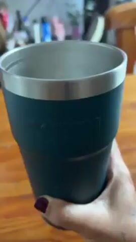 A hand with dark red nail polish and a tattoo holds a dark blue and silver insulated cup, revealing a lemon slice at the bottom of the cup.