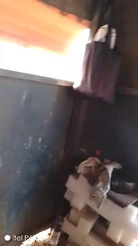 Here is a description of the video:

The video shows the inside of a dark space, possibly a truck or storage container. There is a bag hanging, some items stacked on the floor, and a person's legs are visible at the bottom. The camera moves, and a man in a security guard uniform looks in. The camera then looks out the opening of the space, showing an outdoor area with shipping containers.