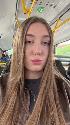 The video is a selfie taken from the perspective of a young woman on a bus. She has long, straight blonde hair and is wearing a dark grey t-shirt and a brown jacket. A silver necklace with a cross pendant is visible. Her lips are slightly pursed. The background shows the interior of a bus with yellow and silver grab handles, seats, and windows. Other passengers are faintly visible in the background. The lighting suggests it is daytime.