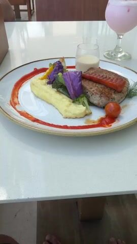 The video shows a plate of food and a pink drink on a white table. The plate contains a piece of grilled tuna topped with a slice of salmon, mashed potatoes, broccoli, purple cabbage, cauliflower, a cherry tomato, and a small glass of white sauce. There are red sauce decorations around the plate. The drink is in a stemmed glass with a straw. The table is on a patio with brown chairs and plants in the background. The camera zooms in slightly on the food. Parts of feet are visible at the bottom of the screen.