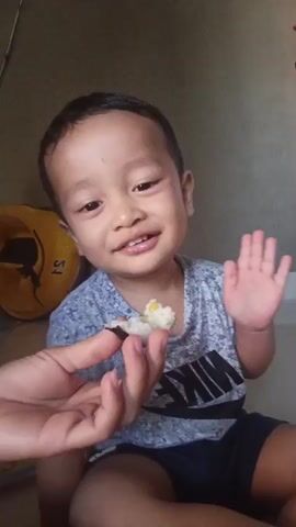 A young child sits on the floor in front of a camera. The child is wearing a grey t-shirt with a black Nike logo and dark blue shorts. The child is smiling and waving at the camera. A hand enters the frame from the left side of the screen, holding a piece of food, which appears to be a sushi roll or similar item. The child opens their mouth and takes a bite of the food. The background is a plain wall and a white shelf with a red object on top. The lighting is natural.