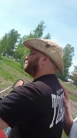 The video depicts a man and a woman in an outdoor setting. Initially, the woman, wearing a straw hat, stands to the right of the man, who has a beard, wears a black T-shirt with white letters on the back, and a hat. The woman places her hand on his back. They appear to be outdoors near a house under a clear sky. The scene shifts to the man on a red motorcycle. He turns his head to look at the camera. The background includes trees and a grassy field. The camera then refocuses on the man's back.