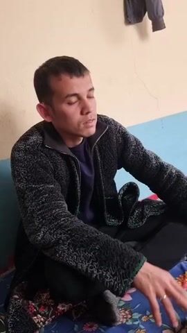 The video shows a man sitting cross-legged on a bed with a floral pattern. He is wearing a black jacket with a textured pattern, a purple shirt, black pants, and grey socks. He has short, dark hair and a ring on his finger. The background consists of a light yellow wall, a blue panel behind the bed, and a piece of clothing hanging from the wall. The man raises his hand and then looks to his right.