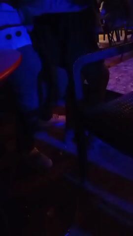 The scene is dark, illuminated by colored lights, possibly in an outdoor bar or club at night.  People are seen sitting at tables; their bodies are mostly out of view. A red table is visible with a water bottle.  The floor is paved with what appears to be cobblestones or tiles in a patterned design. People are standing and walking around in the background. The camera angle is low, looking upwards. The overall atmosphere is lively and social.