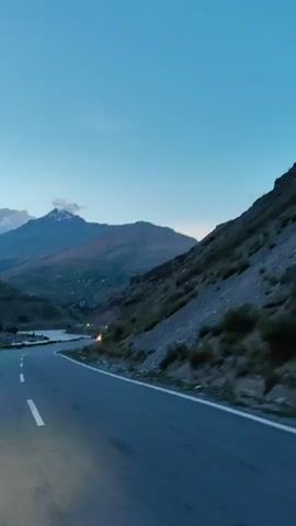 The video shows a winding road through a mountainous landscape at dusk. The sky is a clear, pale blue, with some wispy clouds near the peaks of the mountains. The mountains themselves are rugged and steep, with some vegetation on the slopes. The road is paved and has white lane markings. There is a single headlight visible in the distance, suggesting another vehicle is on the road. The overall impression is one of solitude and natural beauty.