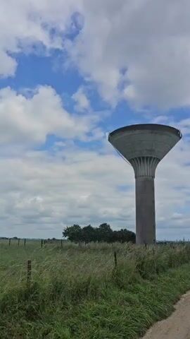 The video shows a water tower in a rural setting. The tower is tall and cylindrical with a large bowl-shaped top. It is surrounded by a grassy field with a dirt road leading away from it. The sky is blue with scattered white clouds. There are a few trees in the distance. The camera is moving slowly, panning across the landscape. The overall impression is one of tranquility and openness.